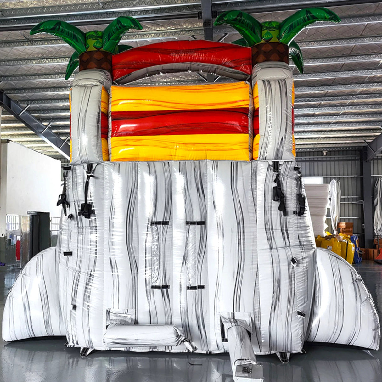 15ft Fire Island Commercial Single Lane Inflatable Water Slide featuring tropical palm trees, red curved slide, yellow inflatable base, and extended splash pool in warehouse setting