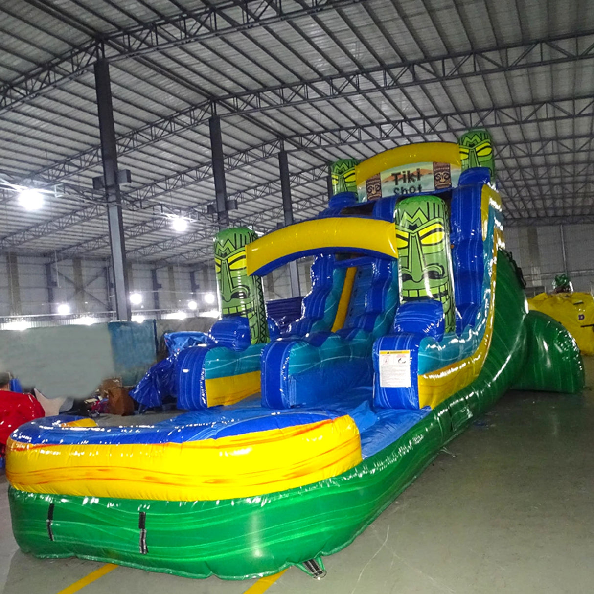 Side view of Tiki Shot single lane water slide in indoor facility, showcasing its length, blue sliding lane, tiki mask decorations, and extended green and yellow splash pool area.