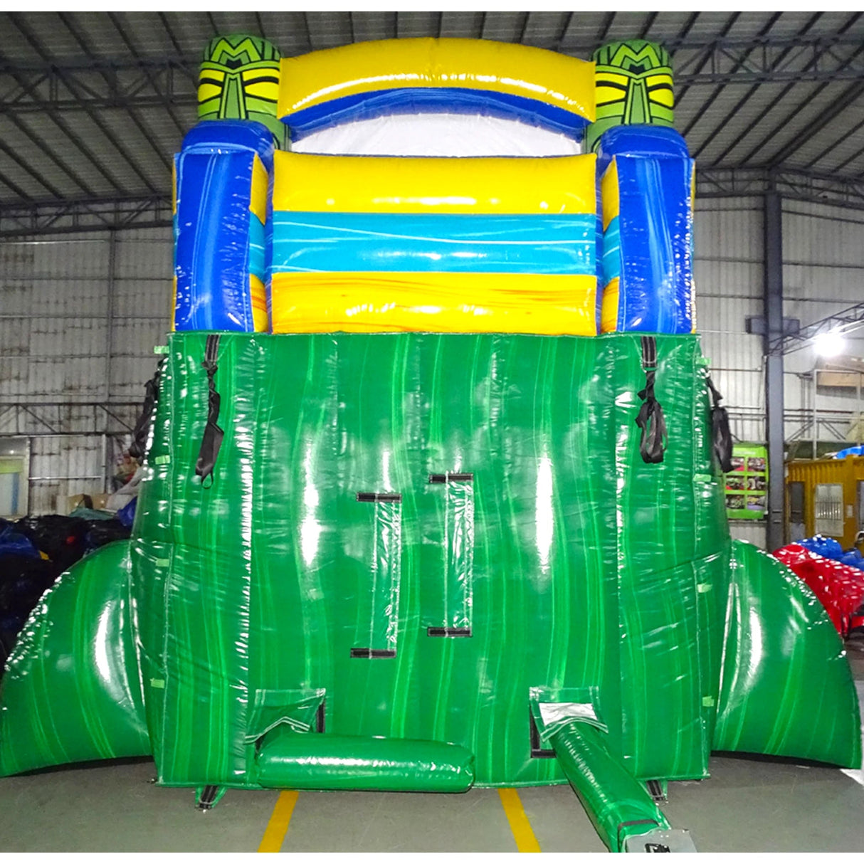 Rear view of Tiki Shot inflatable water slide, displaying tall green structure with climbing area, safety features, and tropical-themed top section with tiki masks and yellow accents.