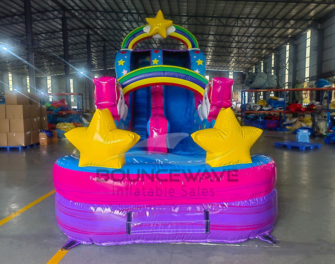 Colorful inflatable water slide with unicorn theme featuring rainbow arches, star decorations, and yellow star-shaped pool floats in indoor warehouse setting.