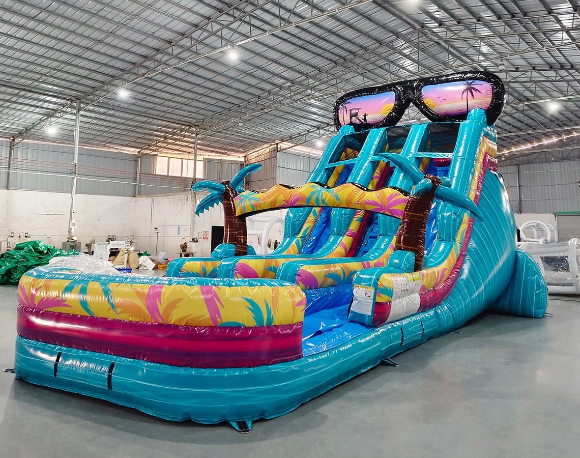 Side view of 18ft Island Flow water slide showing vibrant blue, yellow, and pink colors, inflatable palm trees, giant sunglasses decoration, and a large splash pool area, displayed in a spacious indoor facility with metal roofing.