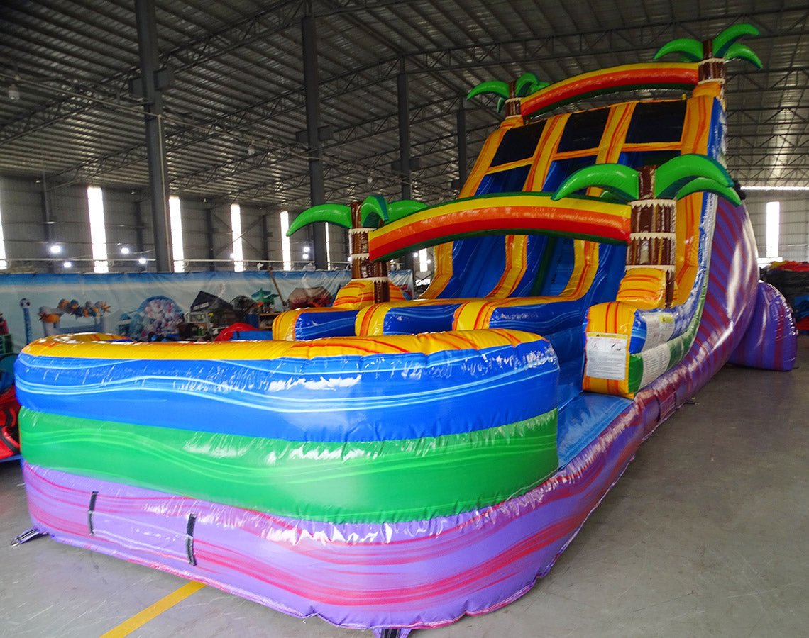 Side view of the 18ft Goombay Splash Center Climb water slide, showcasing its three blue sliding lanes, yellow borders, inflatable palm trees, rainbow arch, and large multicolored splash pool area, set up in a spacious indoor warehouse.