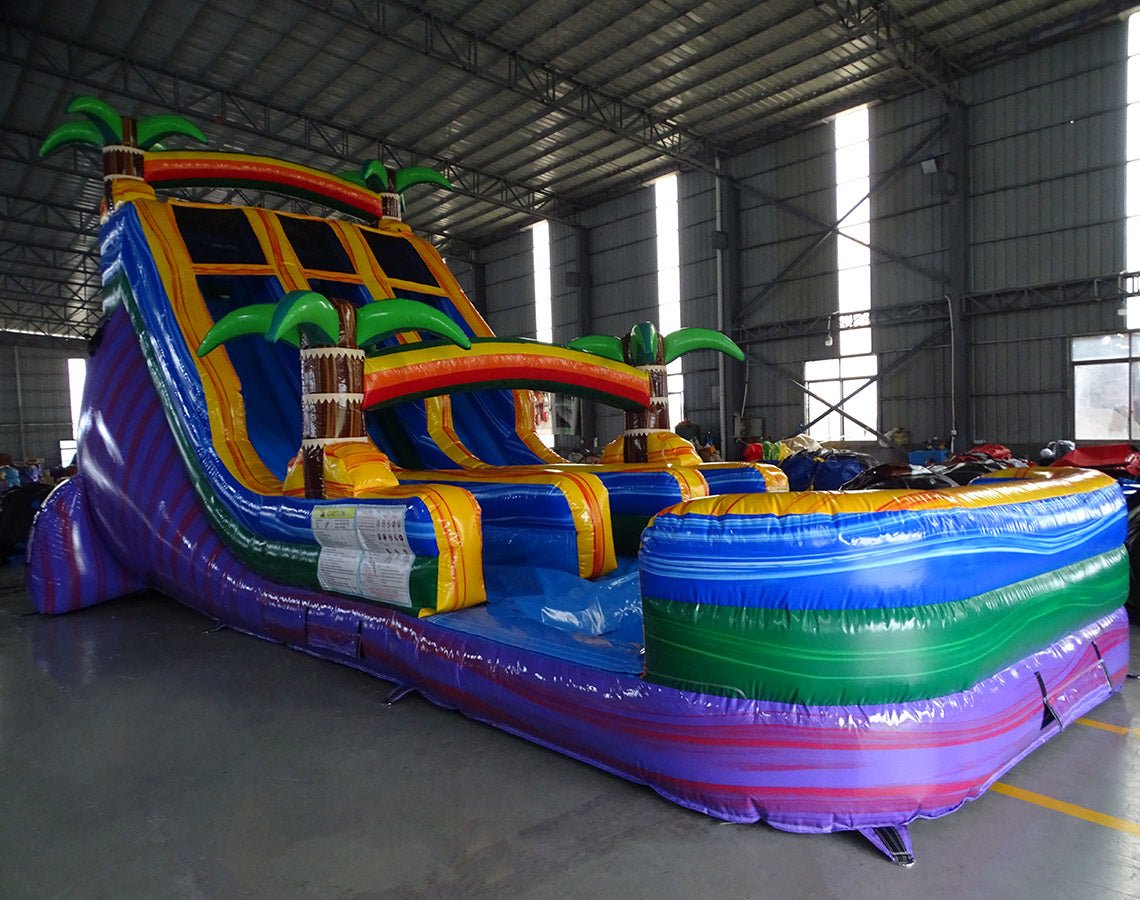 Angled view of 18ft Goombay Splash inflatable water slide featuring blue sliding lanes, yellow borders, palm tree decorations, rainbow arch, and a colorful splash pool, displayed in a large indoor facility with metal roofing visible.