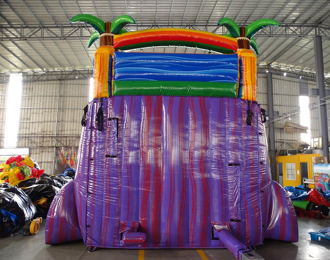 Rear view of 18ft Goombay Splash water slide showing vibrant purple and red base, green and blue top sections, inflatable palm trees, and rainbow arch, displayed in an indoor facility with other inflatables visible.