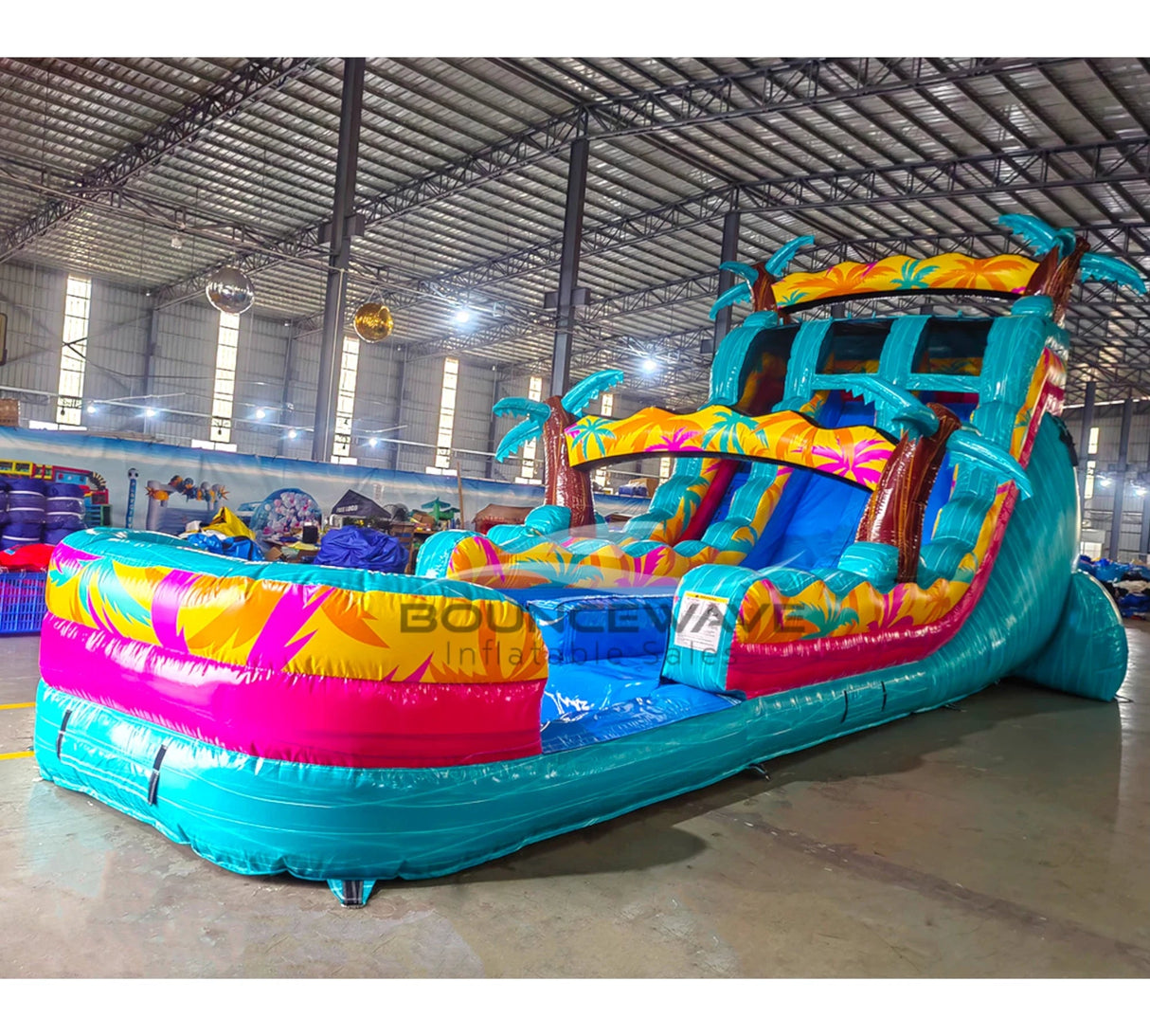 Side view of Island Flow Palms inflatable water slide showing full length with dual lanes, palm trees, and extended splash pool in warehouse environment.