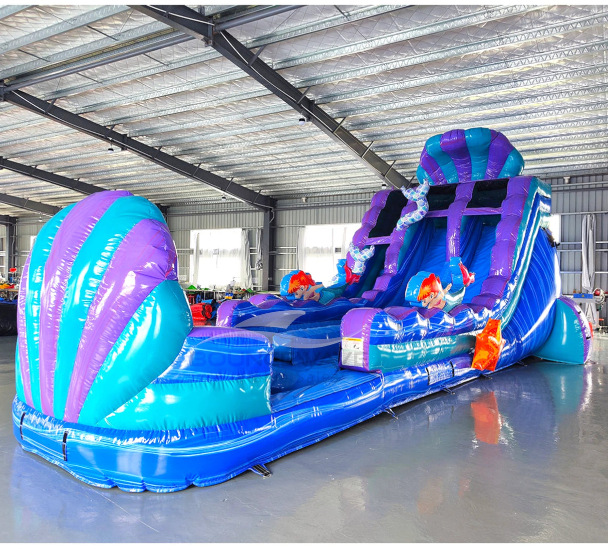 Large inflatable water slide with mermaid theme, featuring blue and purple colors, dual lanes, and shell-shaped top in indoor warehouse setting.