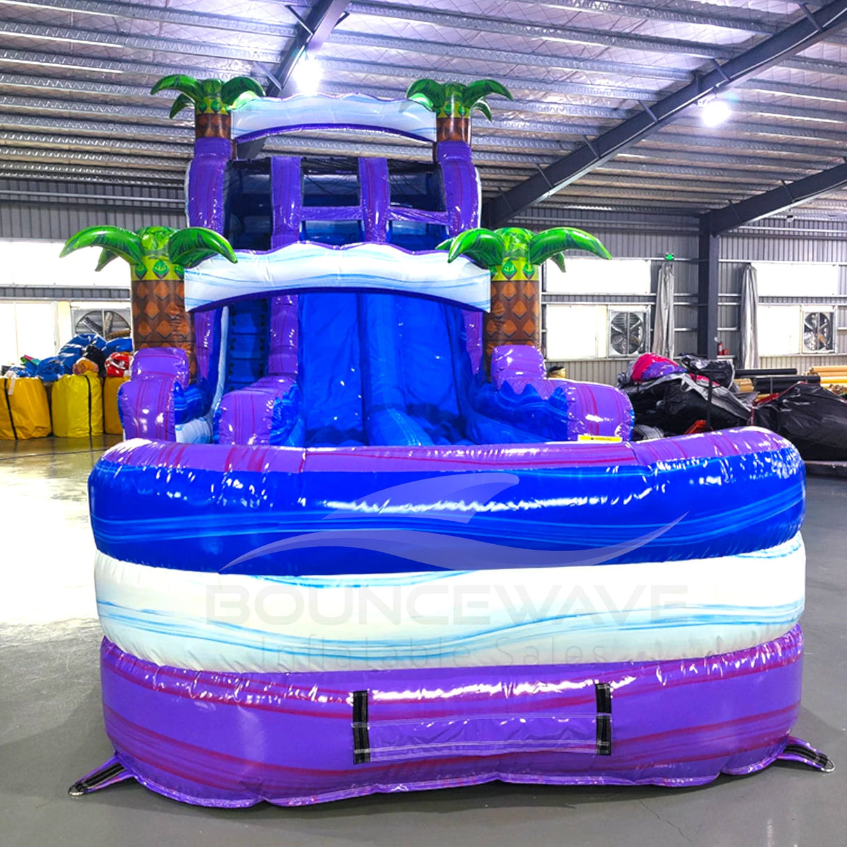 Front view of Purple Thunder hybrid water slide in indoor warehouse, showing tall structure with dual purple sliding lanes, palm trees, and large blue splash area.