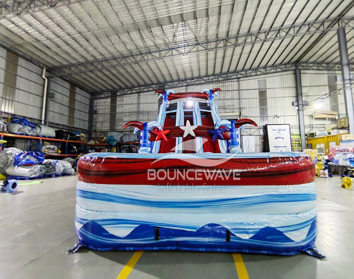 Front view of 18ft Star Spangled Splash Center Climb water slide featuring patriotic color scheme, star decorations, three sliding lanes, and a spacious splash pool with red, white, and blue wave patterns, set up in an indoor warehouse surrounded by other inflatables and equipment.