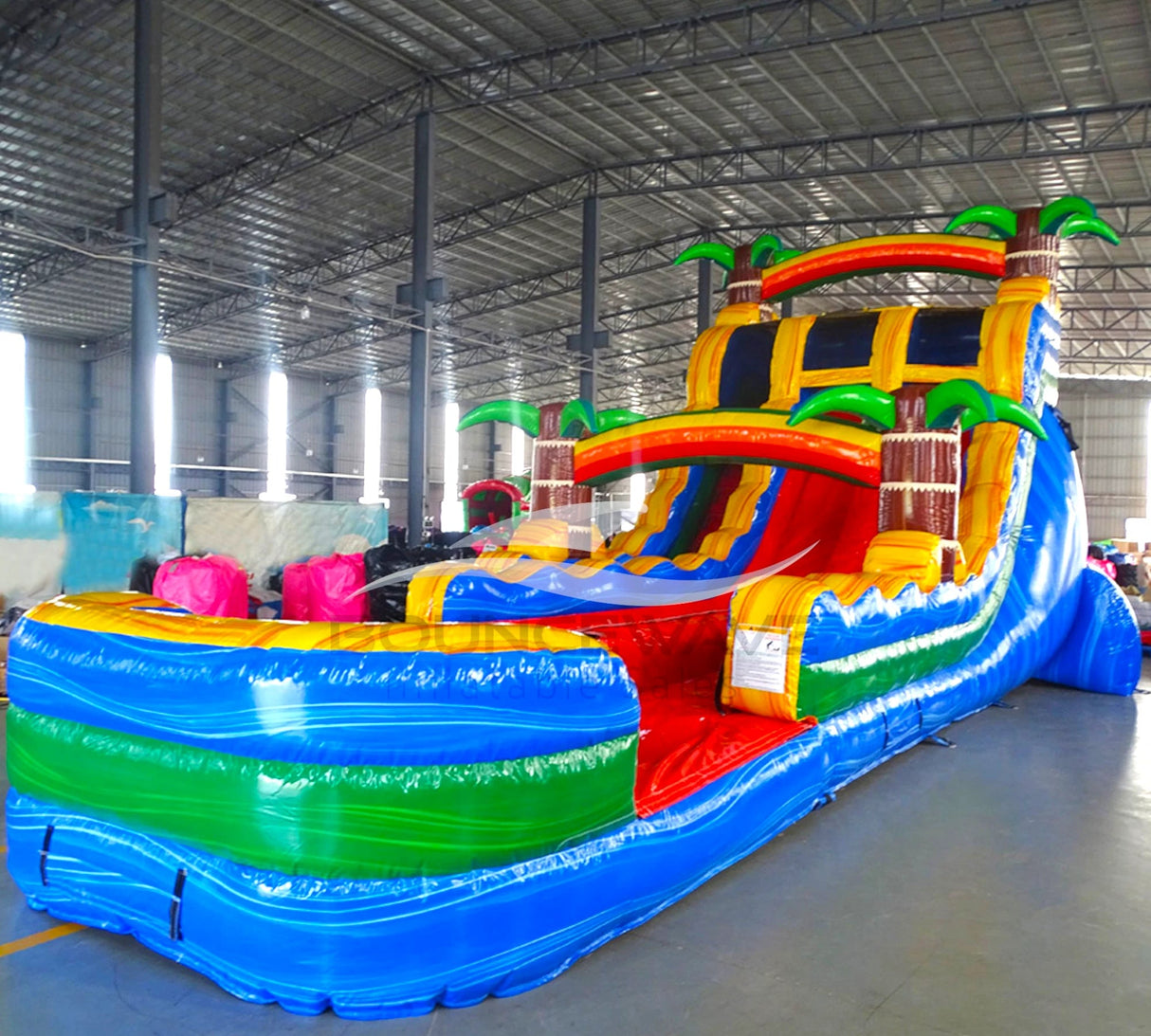 Side view of Tropical Oasis hybrid water slide in indoor warehouse, showing full length with dual sliding lanes, palm trees, and large splash area with blue and green colors.