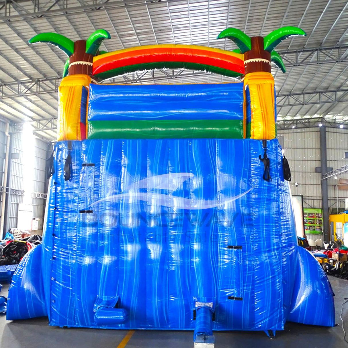 Rear view of Tropical Oasis inflatable water slide, displaying tall blue structure with climbing area, safety features, and tropical-themed top section with palm trees and rainbow arch.