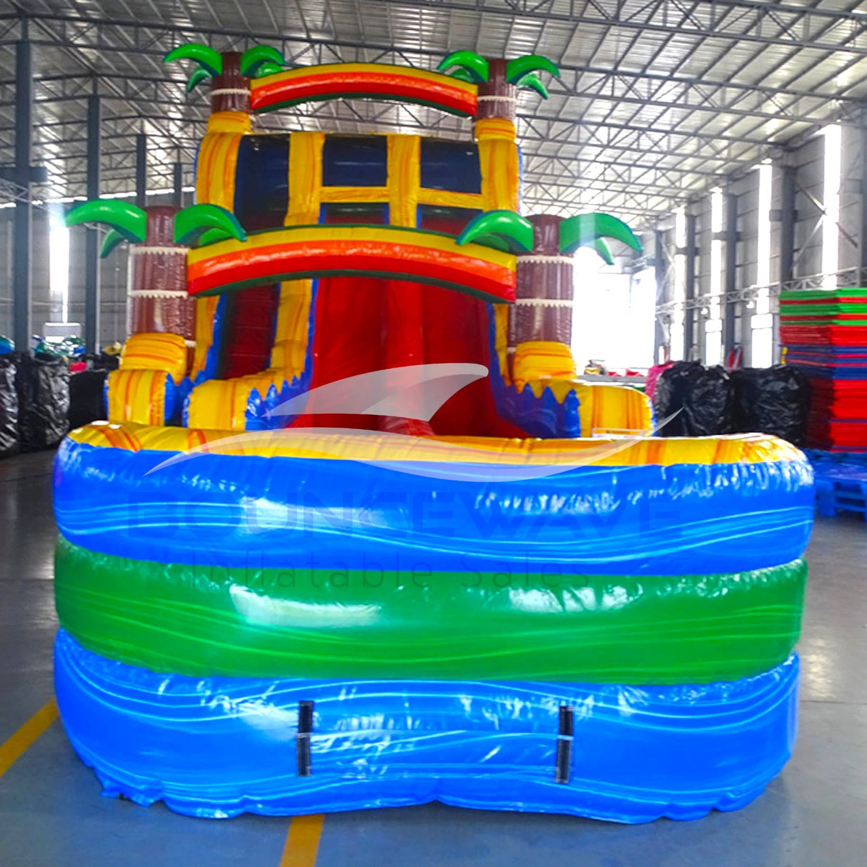 Front view of Tropical Oasis hybrid water slide in indoor facility, showcasing its height, dual lanes, palm tree decorations, and extended blue and green splash pool area with wave-like design.