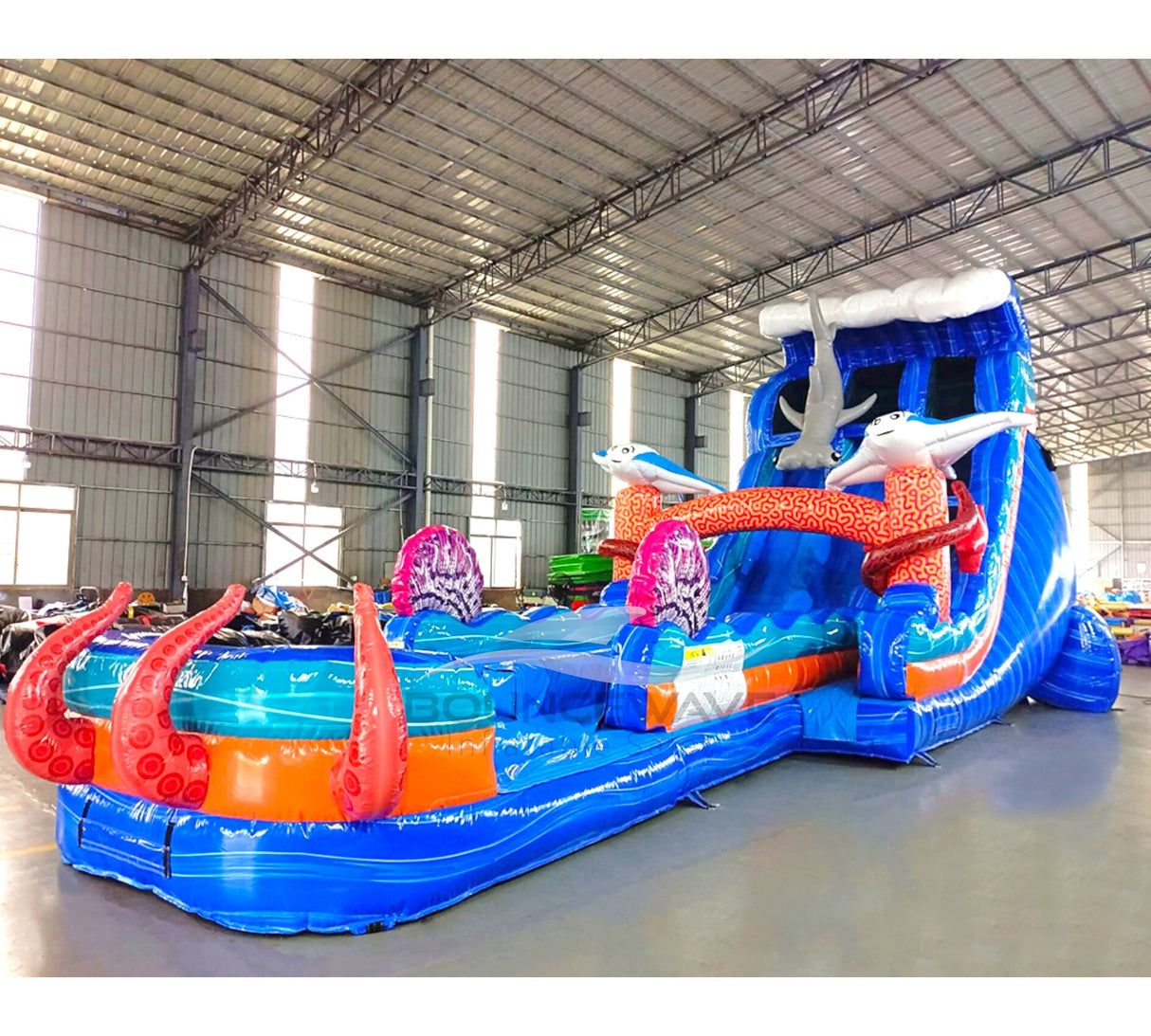 Side view of 20ft Hammerhead Reef Hybrid DL water slide showing vibrant blue colors, inflatable shark figures, octopus tentacles, and a large splash pool area. The slide features both open and enclosed sliding sections, displayed in an indoor facility with other inflatables visible.