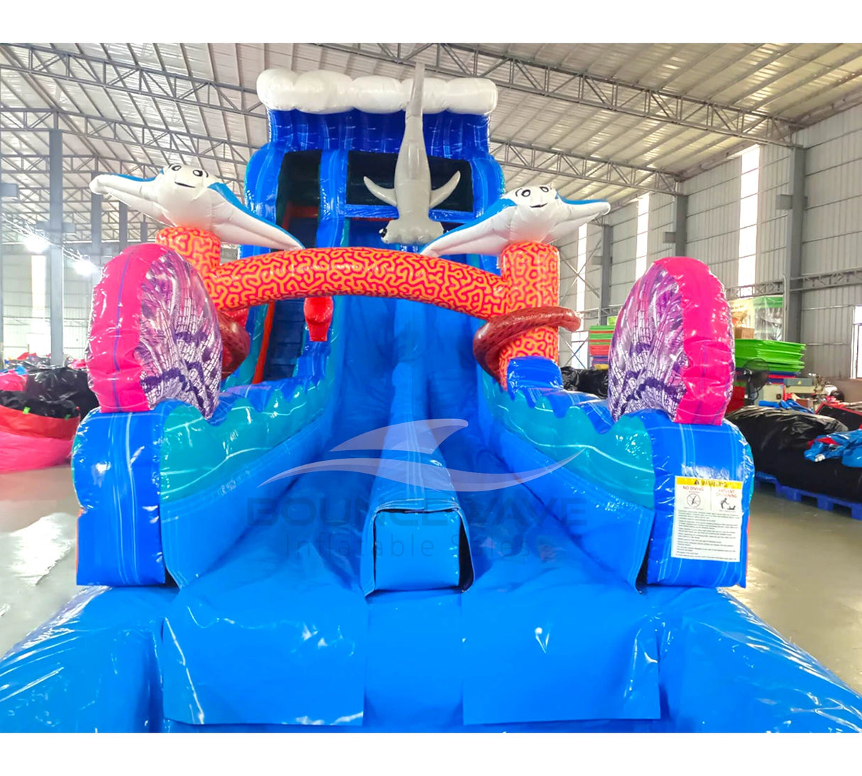 Front view of the 20ft Hammerhead Reef Hybrid DL inflatable water slide, showcasing its blue sliding lanes, shark decorations, octopus tentacles, and spacious splash pool. The slide combines open and tube slide elements, set up in an indoor warehouse with other inflatables visible in the background.