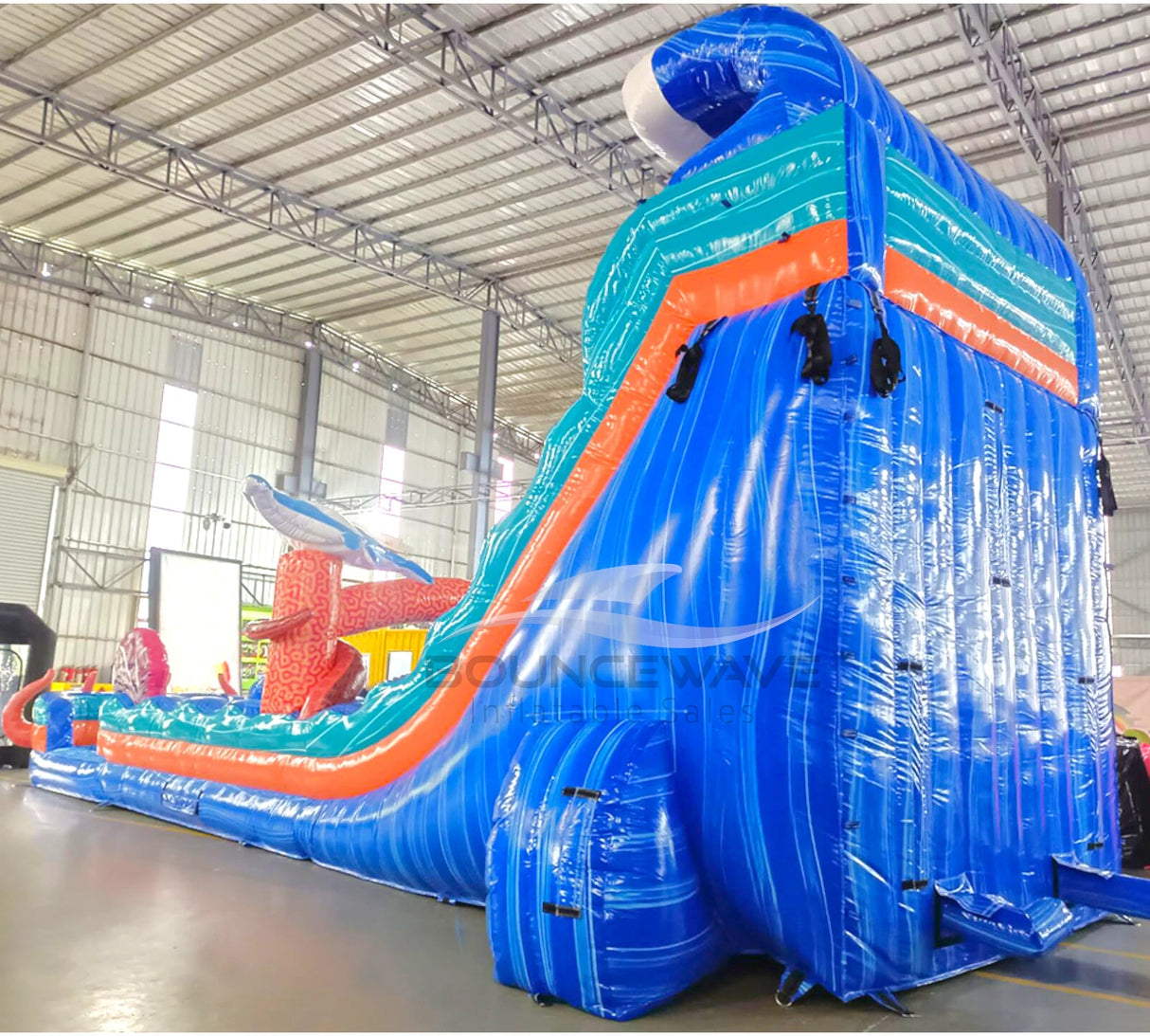 Large inflatable water slide with ocean theme, featuring blue sliding lanes, shark decorations, and a spacious splash pool area with octopus tentacles. The slide combines open and enclosed sections, set up in an indoor warehouse with metal roofing visible.