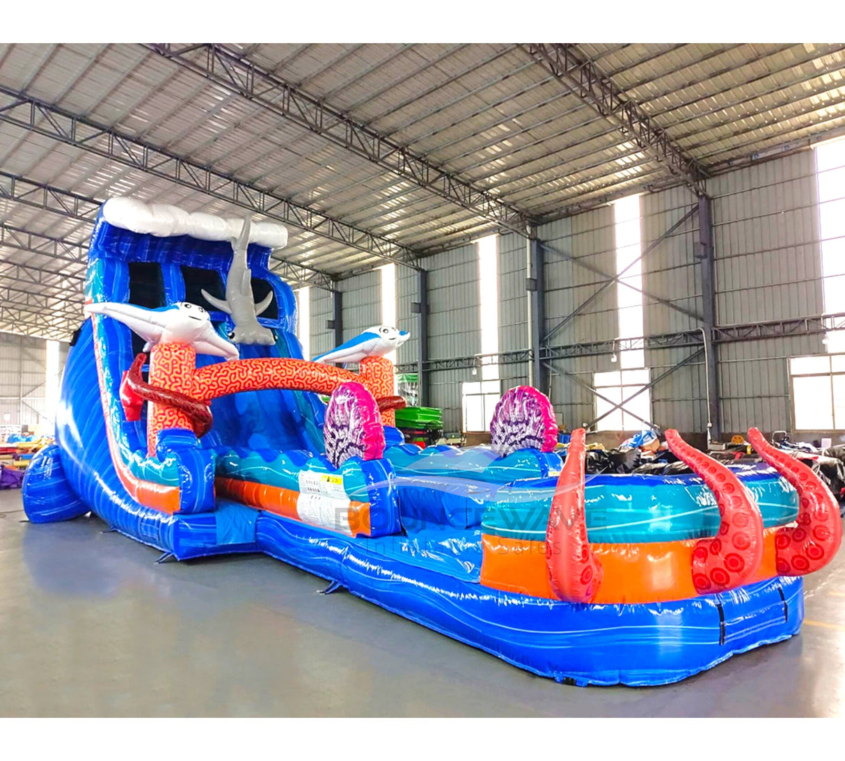Large inflatable water slide with ocean theme, featuring blue sliding lanes, shark decorations, and a spacious splash pool area with octopus tentacles. The slide combines open and enclosed sections, set up in an indoor warehouse with metal roofing visible.