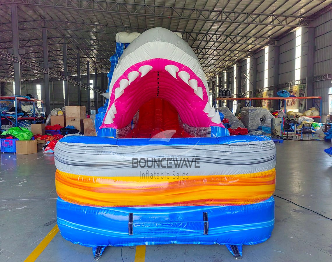 20ft Rip Curl Shark Attack 2pc Hybrid Dual Lane Inflatable Water Slide featuring giant shark mouth entrance, blue water slide, and colorful inflatable base in warehouse