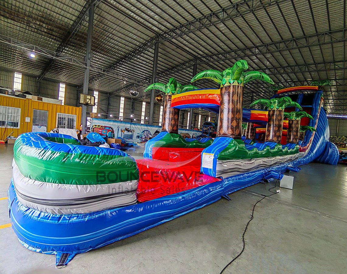 24ft Baja Splash DL 2pc Commercial Water Slide with Inflatable Slip & Slide showcasing inflatable palm trees, blue slide, and extended slip and slide in indoor facility