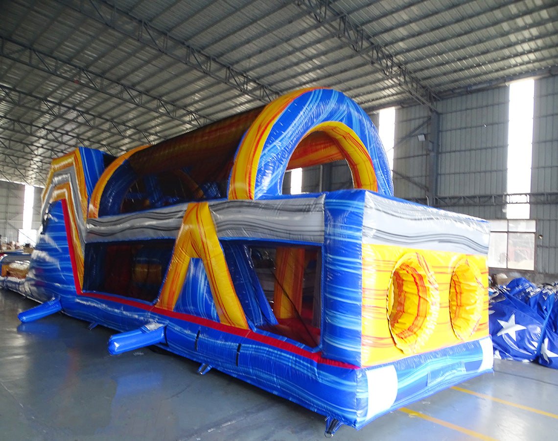 Three-quarter view of 53ft XL Rip Curl Commercial Hybrid Inflatable Obstacle Course displaying its full length. The structure has a blue and yellow design with dual red slides, multiple obstacles, and a large circular splash pool. Set in a large indoor warehouse with metal roofing and other inflatable products visible.