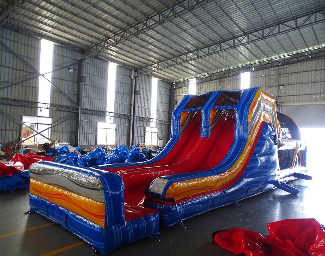 Side view of 53ft XL Rip Curl Commercial Hybrid Inflatable Obstacle Course featuring dual red slides emerging from a blue and yellow structure. The inflatable includes multiple obstacles, tunnels, and a large circular splash pool. Set up in a spacious warehouse with metal roofing and bright lighting visible.