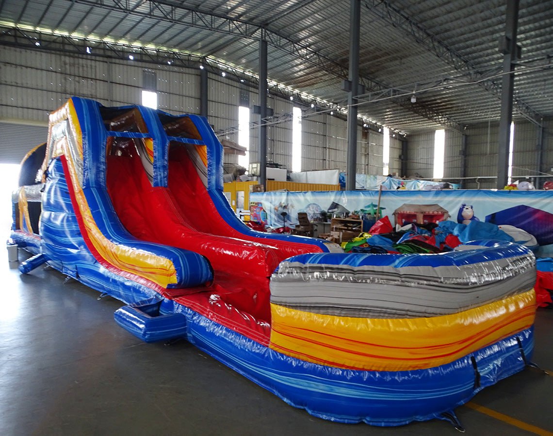 Side view of 53ft XL Rip Curl Commercial Hybrid Inflatable Obstacle Course featuring a blue and yellow color scheme. The inflatable structure includes dual red slides, multiple obstacles, and a large circular splash pool. Set up in a spacious warehouse with metal roofing and bright lighting visible.