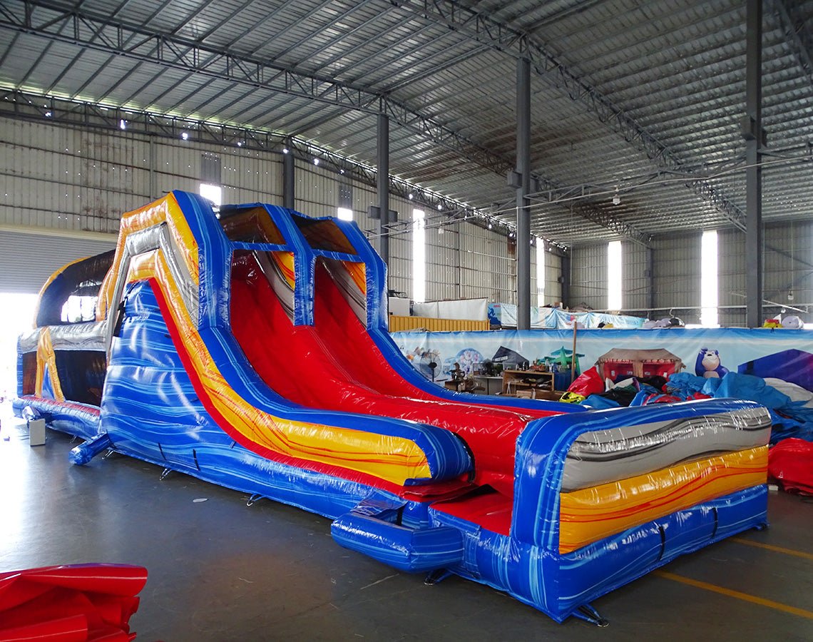 Close-up view of 53ft XL Rip Curl Commercial Hybrid Inflatable Obstacle Course entrance area. Features a blue and yellow color scheme with red accents, dual circular entrances, and a large arched top with wave designs. Safety instructions are visible on the front panel. Set up in a warehouse with other inflatable products in the background.