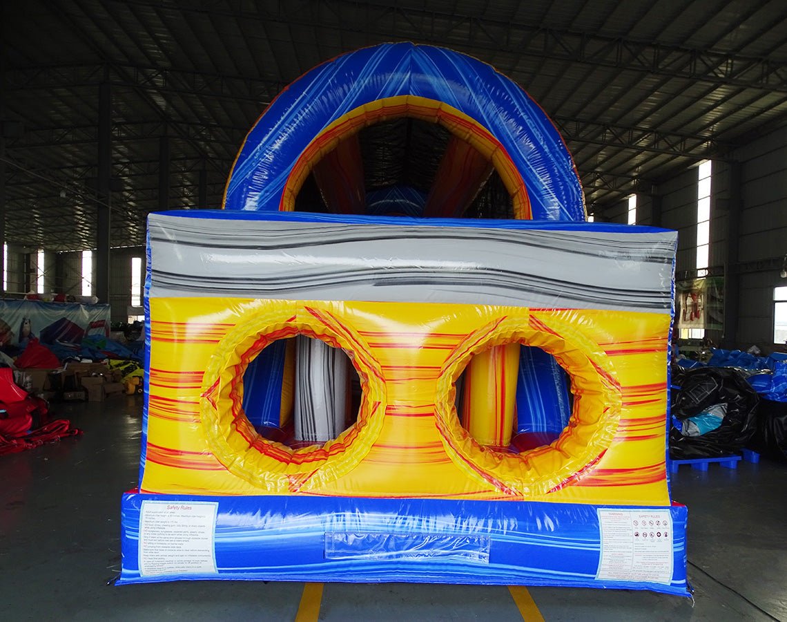 Three-quarter rear view of 53ft XL Rip Curl Commercial Hybrid Inflatable Obstacle Course displaying its full length. The structure has a blue and yellow design with dual red slides, multiple obstacles, and a large circular splash pool. Various other inflatable products and warehouse equipment are visible, showcasing its size and commercial setting.