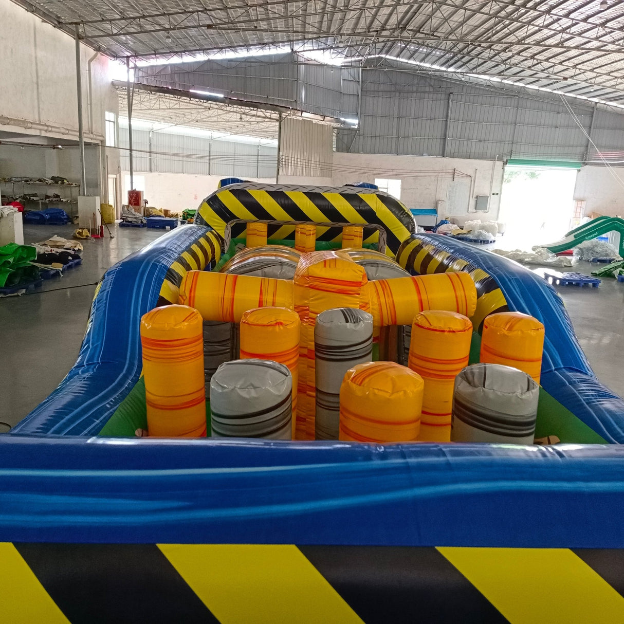92ft Radioactive Run 3-Piece Wet/Dry Inflatable Obstacle Course featuring yellow and black hazard stripes, inflatable barrels, and blue base in warehouse setting