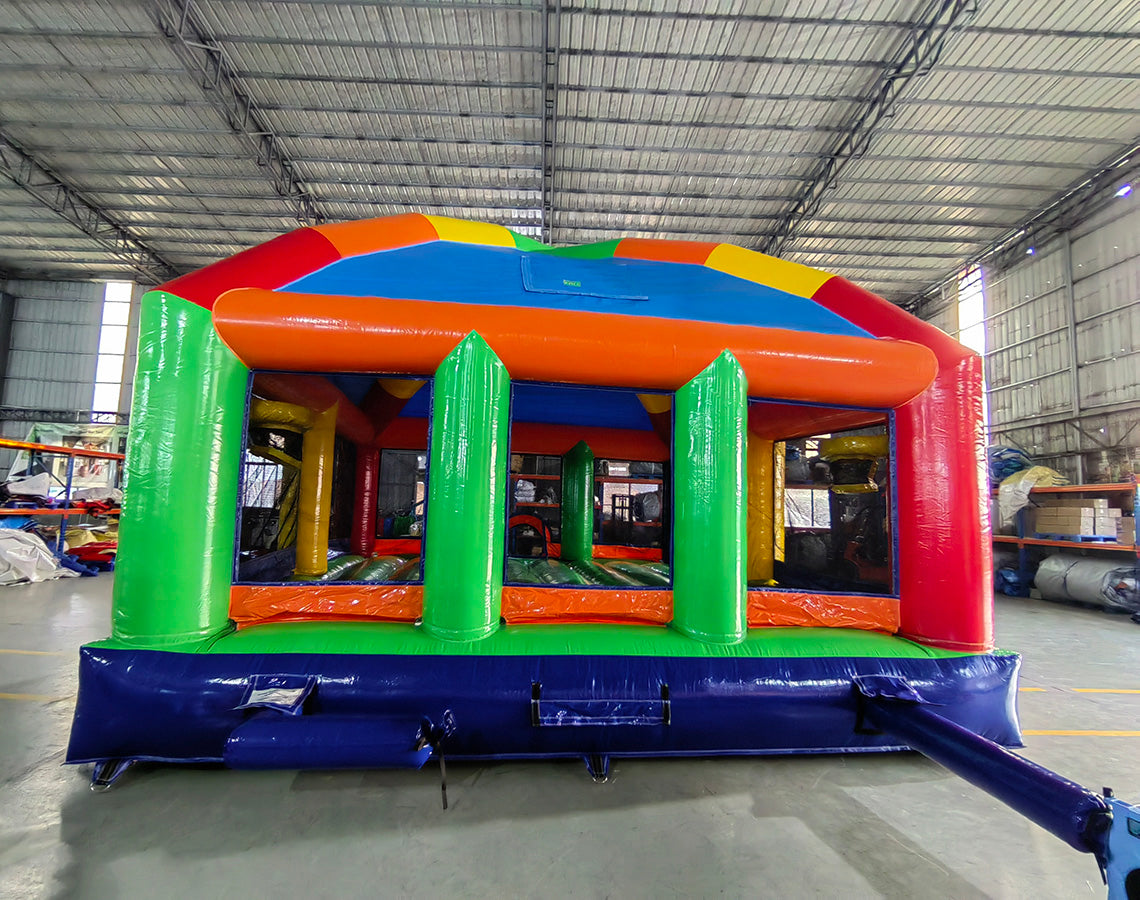 Wide-angle view of Mega Fun Dome Commercial Grade Bounce House in a spacious indoor facility. The inflatable features a rounded blue top with colorful panels, orange support beams, and green pillars. The entrance has a small orange archway. The image showcases the bounce house's size relative to the warehouse, with metal roofing, shelving units, and other inflatable products visible in the background.