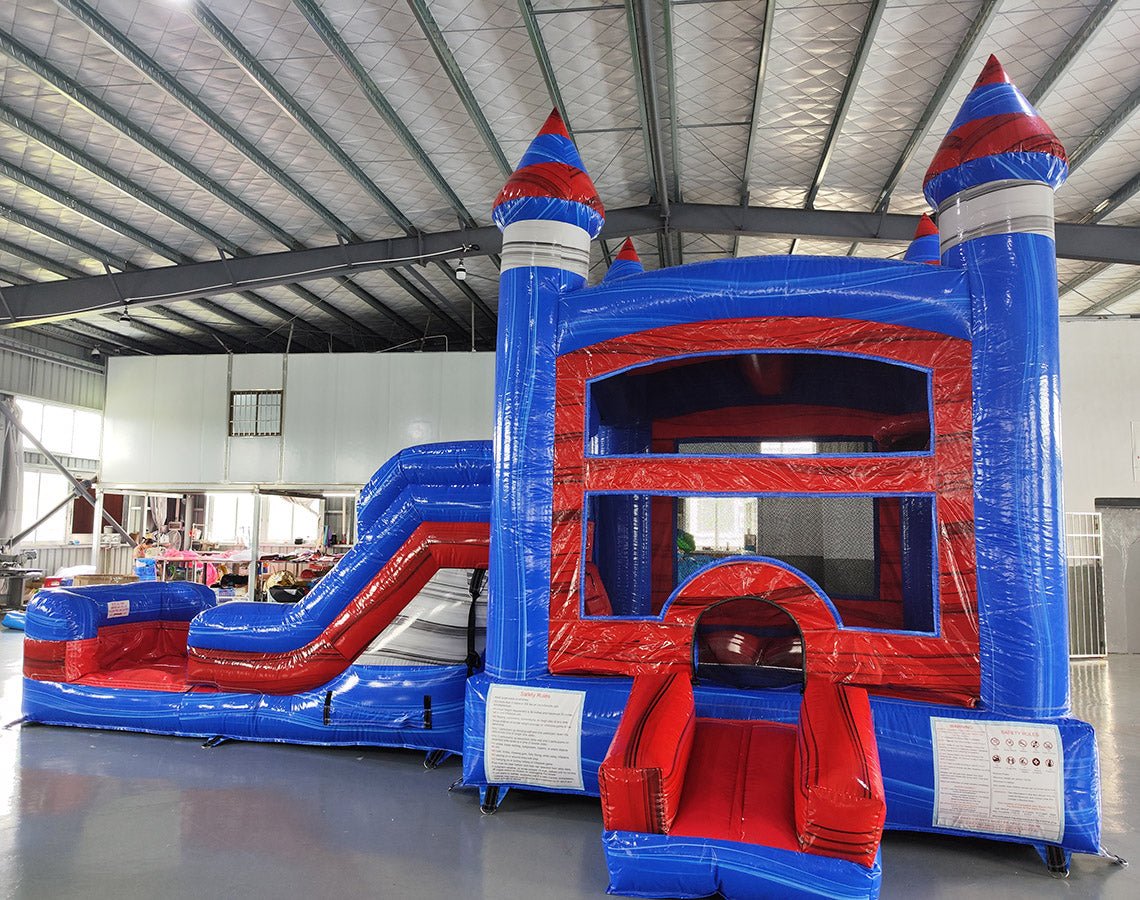Front view of Baja Castle Econo Combo, showing bounce house entrance, slide, and splash pool. Features blue and red castle design with safety instructions visible. Set up in an indoor warehouse with other inflatables and equipment in the background.