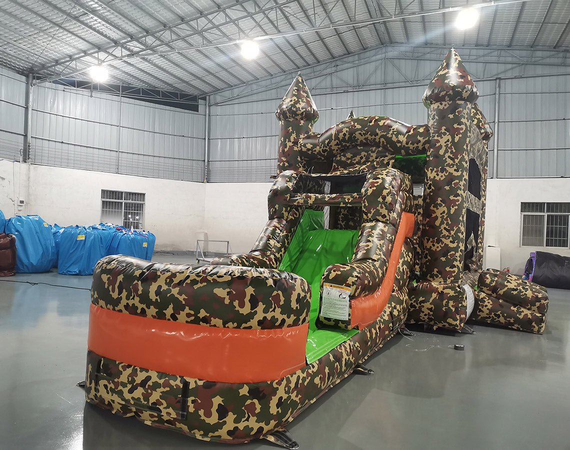 Inflatable military-themed combo featuring a bounce house with camouflage pattern, attached slide, and splash pool. The structure has green, brown, and tan colors with two castle-like turrets. Set up in an indoor warehouse with metal roofing visible.
