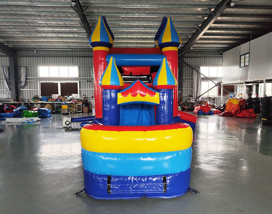Side view of Carnival Econo Combo showing bright red, blue, and yellow design with multiple spired turrets. The combo includes a bounce house, slide, and colorful splash pool area. Set up in an indoor warehouse with other inflatable products and equipment visible.