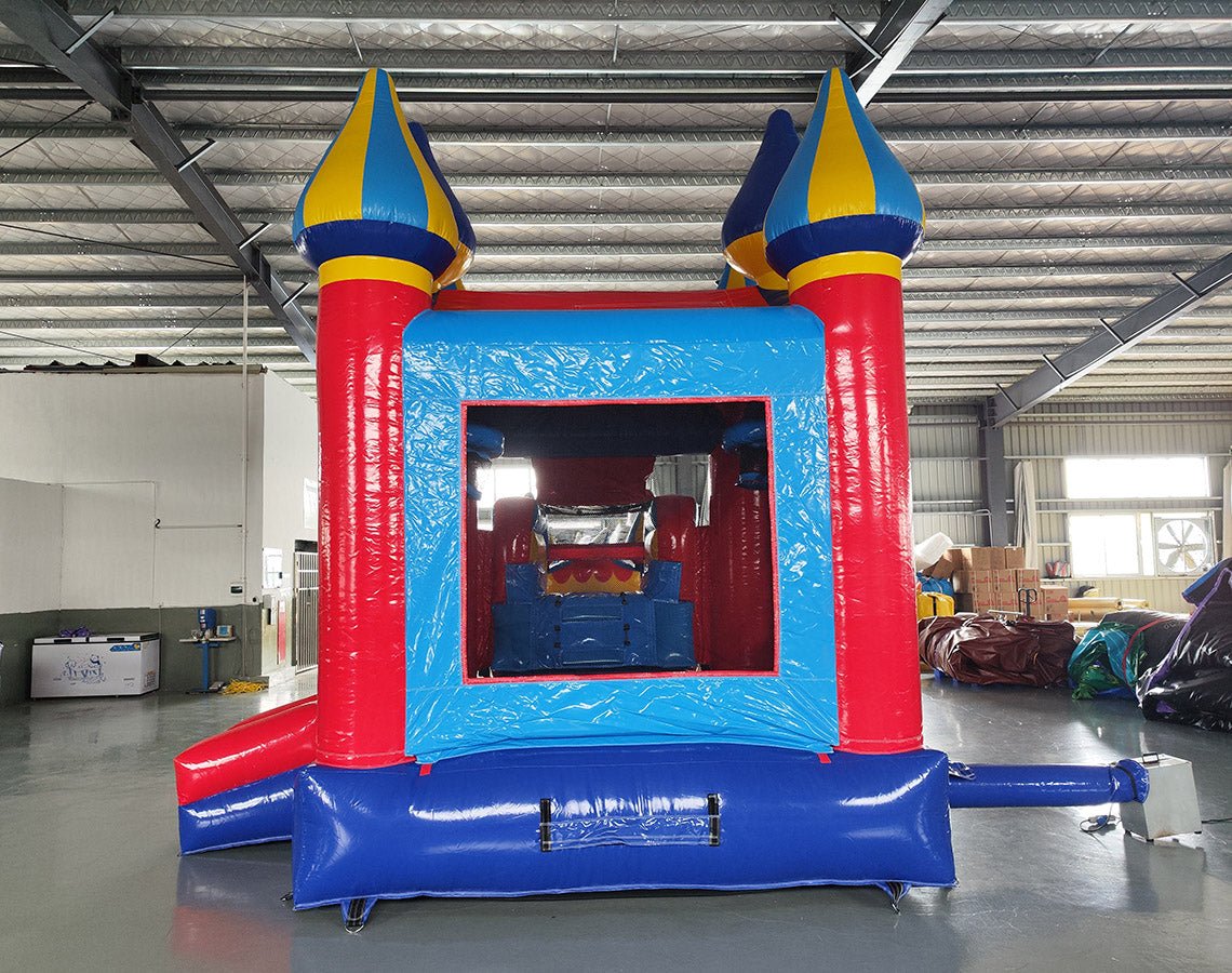 Large inflatable carnival-themed combo featuring a bounce house with colorful spires, attached slide, and splash pool. The structure has vibrant red, blue, and yellow colors with multiple castle-like turrets. Set up in an indoor warehouse with metal roofing and other inflatable products visible in the background.