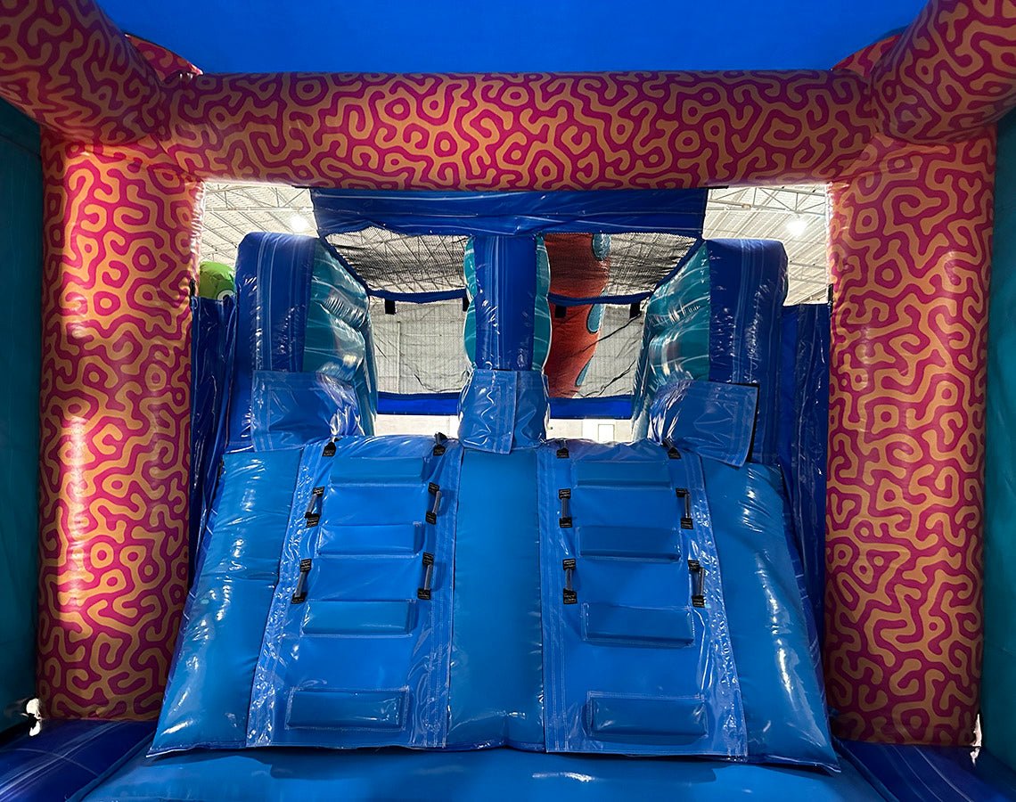 Interior view of Deep Sea Dive Dual Lane Commercial Inflatable Combo (Wet/Dry) showing dual blue slides with safety barriers. The inflatable features an underwater theme with pink and orange coral-like patterns on the walls. Part of the large orange octopus is visible at the top. Set up in a warehouse with metal roofing and support beams visible.