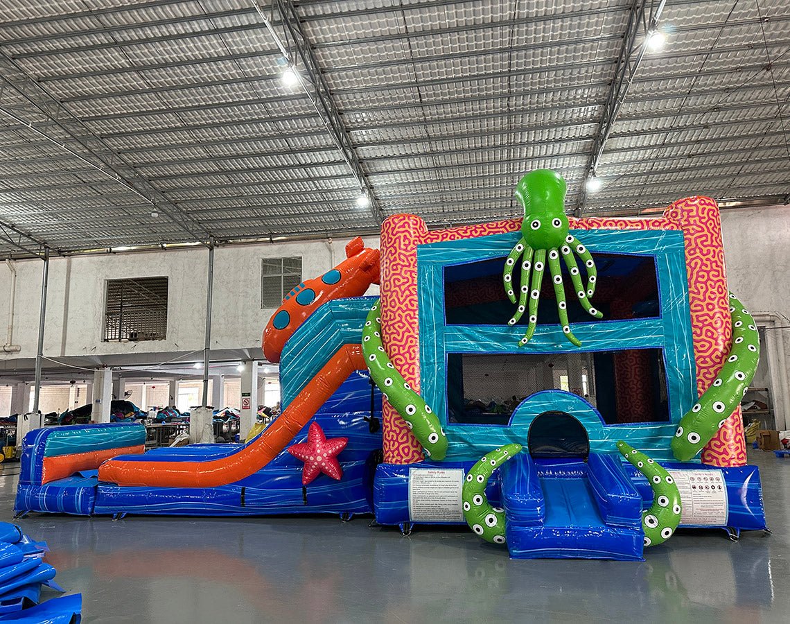 side view of Deep Sea Dive Dual Lane Commercial Inflatable Combo set up in a large indoor facility. Highlights the giant orange octopus, green tentacles, dual blue slides, and blue wave-patterned base with a splash pool. Safety instructions visible on the front. Warehouse interior with metal roofing and various equipment provides scale context.