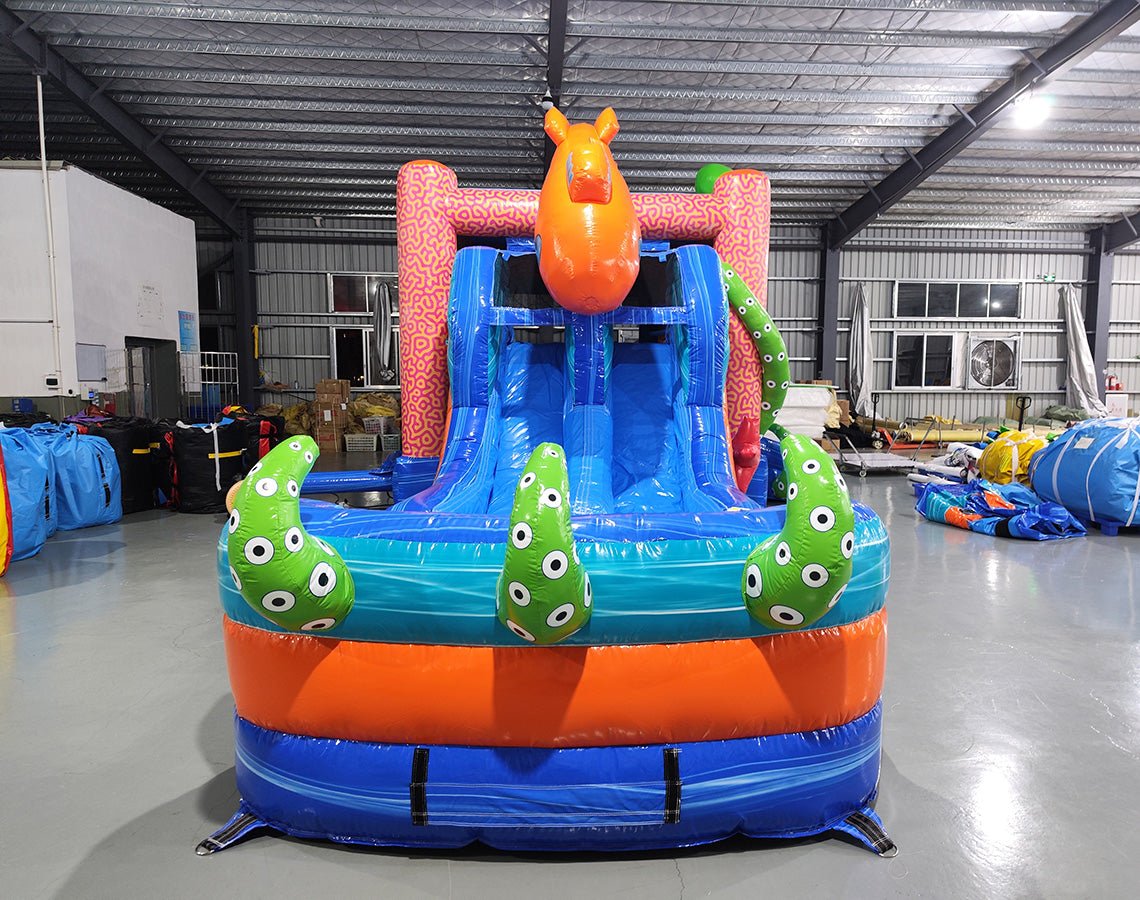 Front view of Deep Sea Dive Dual Lane Commercial Inflatable Combo (Wet/Dry) set up in a large indoor warehouse. Features a giant orange octopus on top, dual blue slides, green tentacles with eyes, and a blue wave-patterned base with a splash pool. Pink and orange coral-like patterns decorate the sides. Various inflatable products and warehouse equipment visible in the background.
