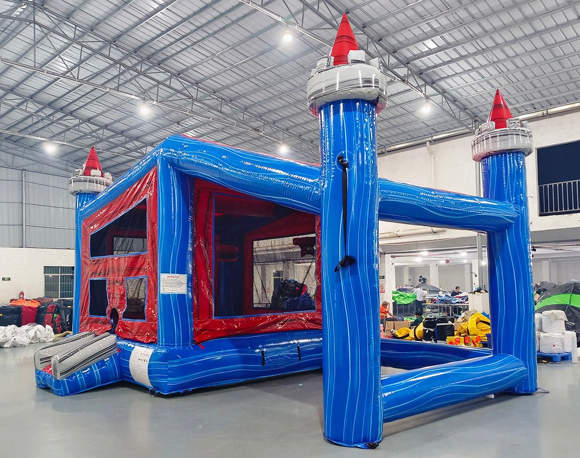 Side angle view of Euro Baja Commercial Grade Canopy Bounce House set up in a large indoor facility. The blue inflatable structure has four tall pillars with white and red castle-like turrets. A red canopy top connects the pillars. The bounce area features red walls with mesh windows. Other inflatable products and warehouse materials are visible in the background.