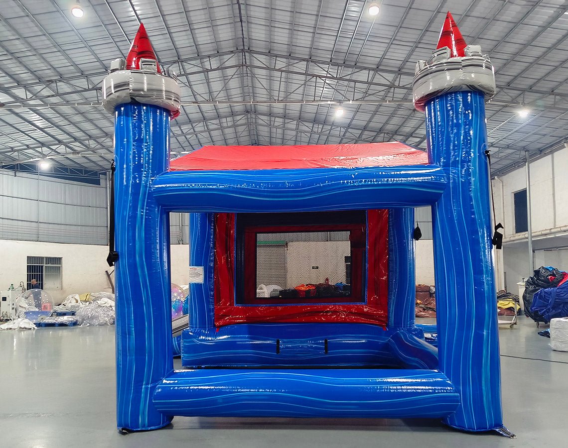Rear view of Euro Baja Commercial Grade Canopy Bounce House in a warehouse environment. Shows the blue inflatable structure with four tall pillars topped by white and red castle-like turrets. A red canopy connects the top of the pillars. The bounce area is visible through the rear opening, showing red interior walls. Various inflatable products and warehouse equipment are seen in the background.