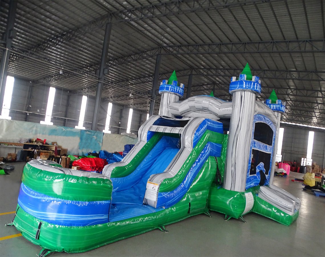 Side view of Euro Green Gush Commercial Econo Inflatable Wet/Dry Combo showing blue and silver castle structure with green turrets, slide, and splash pool. Set up in a spacious indoor warehouse with other inflatable products and equipment visible in the background.