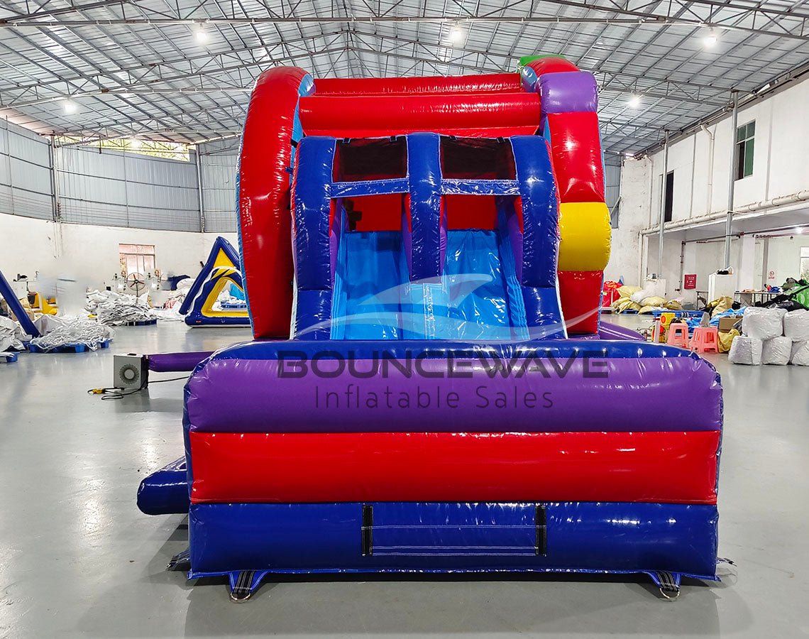 Front view of Ferris Wheel Dual Lane Commercial Inflatable Combo (Wet/Dry) set up in a large indoor facility. Features a red and blue inflatable structure with dual slides and a splash pool area. The "BOUNCEWAVE Inflatable Sales" logo is prominently displayed on the purple base. Other inflatable products and warehouse equipment visible in the background.