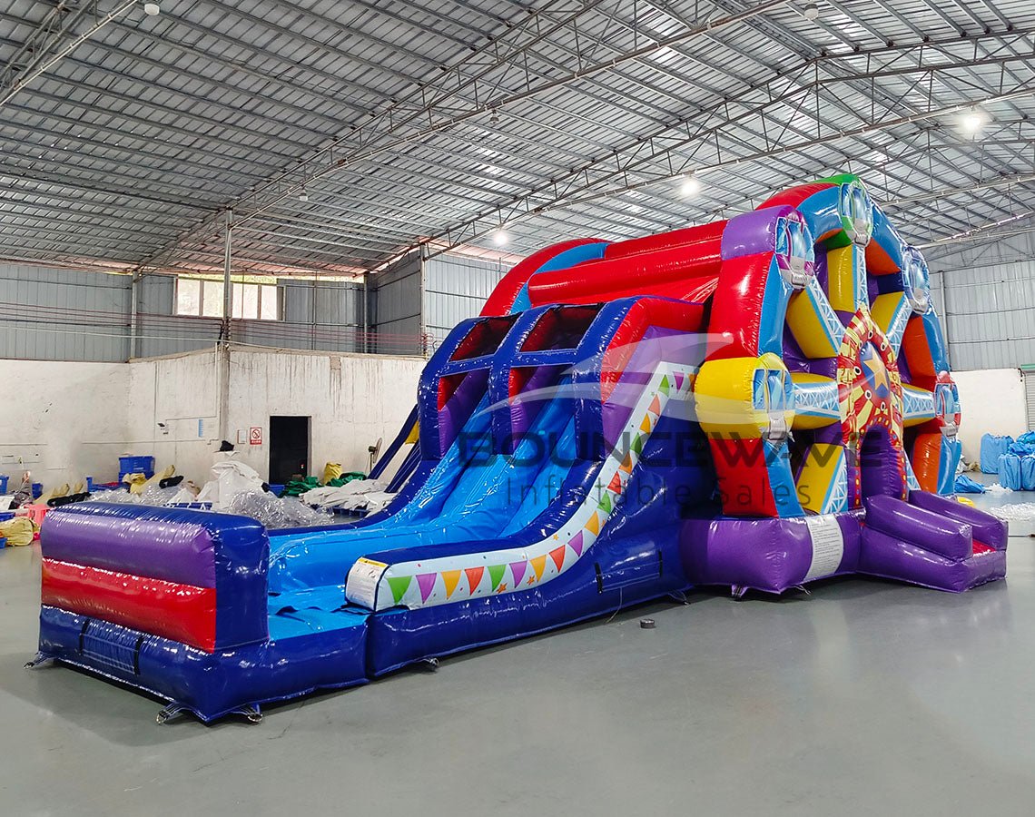 Wide-angle view of Ferris Wheel Dual Lane Commercial Inflatable Combo (Wet/Dry) in a spacious warehouse setting. Showcases the full structure including the Ferris wheel design, dual blue slides, and multicolored base. The warehouse interior is visible with metal roofing, walls, and other inflatable products in the background, providing scale and context for the combo's size.