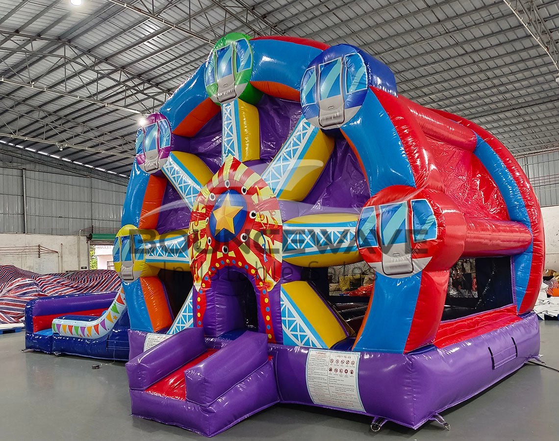 Large colorful inflatable combo featuring a Ferris wheel design with dual slides. The structure has vibrant blue, red, purple, and yellow colors. Central Ferris wheel element has multicolored spokes and a star-shaped center. Two blue slides flank the wheel, leading to a splash pool area. Set up in a spacious indoor warehouse with metal roofing visible.