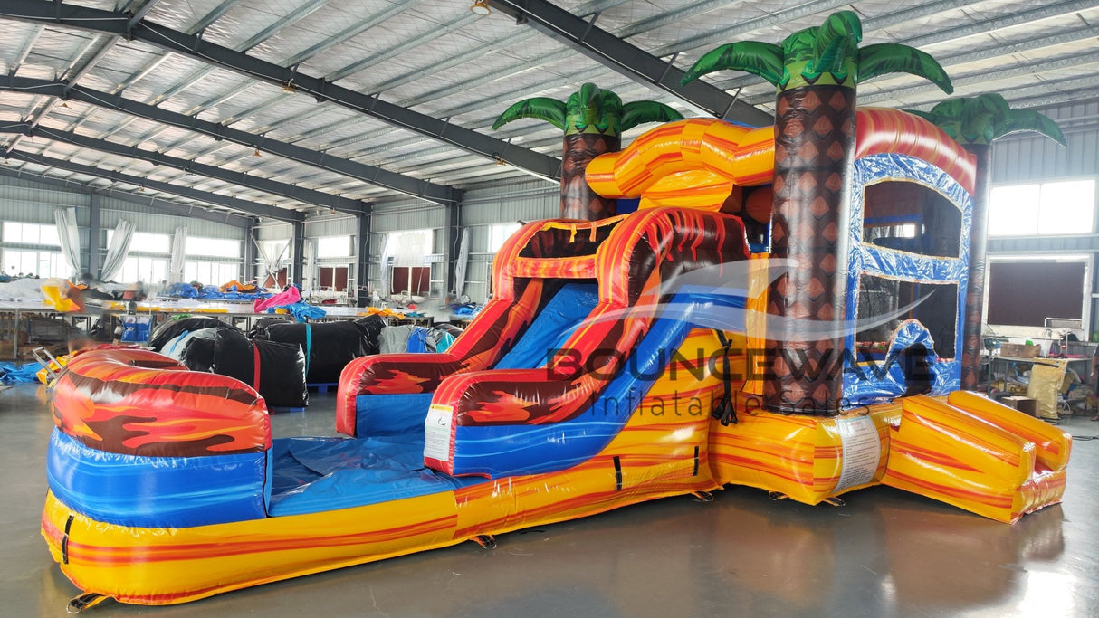Wide-angle view of Fiesta Fire Econo Combo featuring a tropical-themed bounce house with palm tree decorations, curved slide, and large splash pool. The inflatable has bright orange, red, and blue colors and is set up in a spacious indoor warehouse with other equipment and inflatables visible.