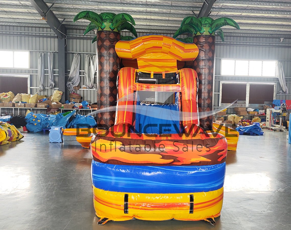 Front view of Fiesta Fire Econo Combo, showcasing the bounce house entrance with palm tree decorations, orange and red slide, and blue splash pool area. Set up in an indoor warehouse with other inflatable products and equipment visible in the background.