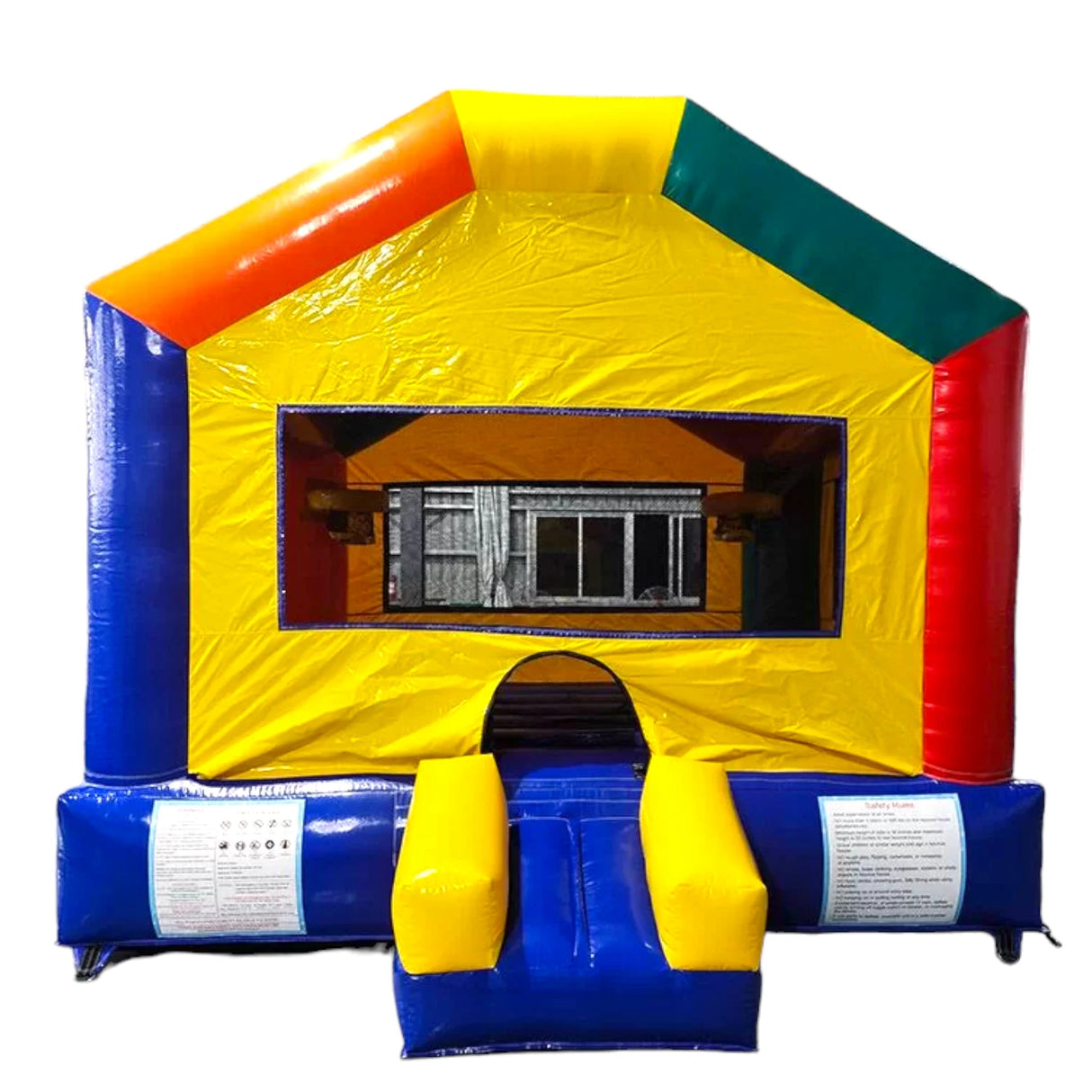 Large inflatable Fun House Commercial Grade Bounce House set up in a spacious indoor warehouse. The structure features a blue base and frame with a red interior visible through mesh windows. Four tall corner pillars topped with white cylindrical turrets and red conical roofs give it a castle-like appearance. The entrance has a blue step and red interior. A canopy extends between the turrets, creating a covered play area. Safety instructions are visible on the front.