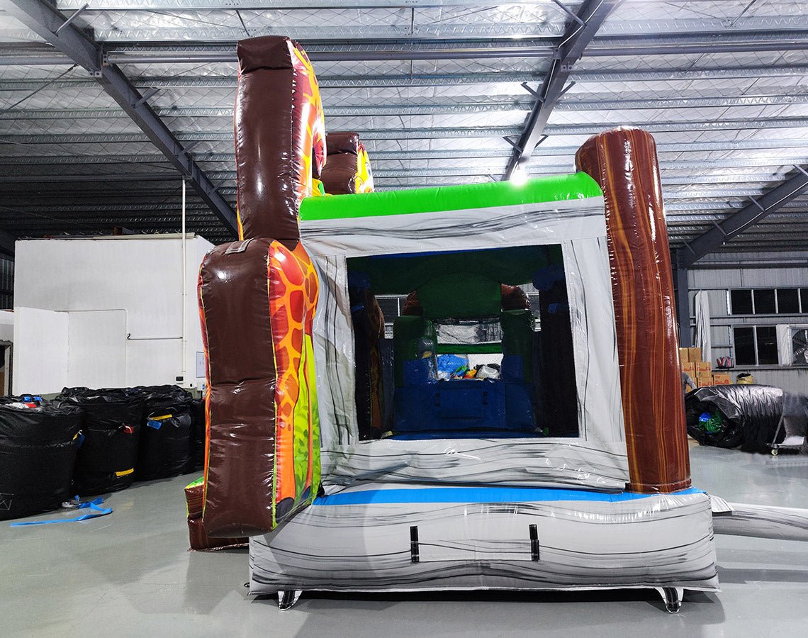 Large inflatable safari-themed combo featuring two tall giraffe figures flanking a bounce house entrance. The structure has vibrant yellow, orange, and green colors with cartoon animal decorations. Includes a slide and splash pool area. Set up in an indoor warehouse with metal roofing visible.