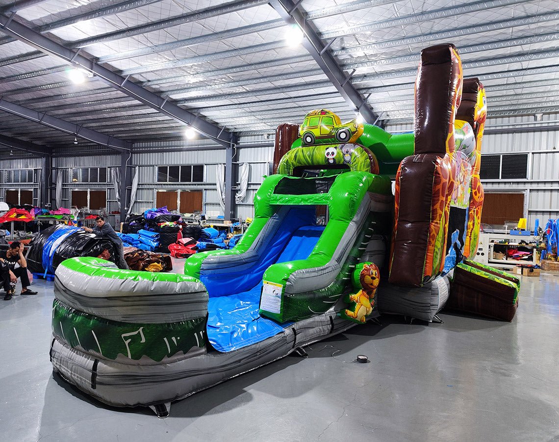 Front view of Giraffe Safari Econo Combo showing two large inflatable giraffes framing a bounce house with colorful animal artwork. Features a blue slide, green obstacles, and a splash pool. Displayed in an indoor facility with other inflatable products visible in the background.