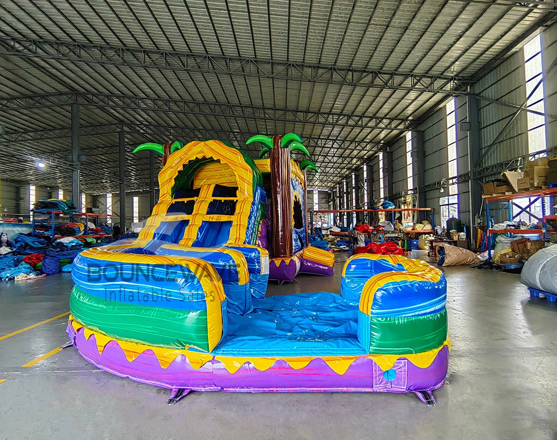 Front angled view of Goombay Curved Dual Lane Commercial Inflatable Combo (Wet/Dry) in a spacious warehouse. Highlights the yellow climbing structure with blue accents, dual blue curved slides, inflatable palm trees, and circular multicolored splash pool. Warehouse interior visible with metal roofing, shelving units, and other inflatable products, providing scale and context for the combo's size.