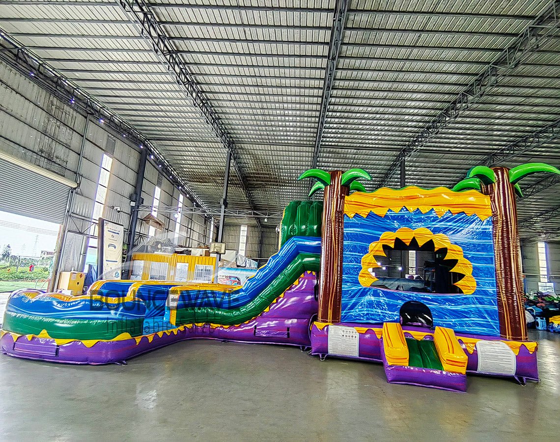 Side view of Goombay Curved Dual Lane Commercial Inflatable Combo (Wet/Dry) in a warehouse setting. Showcases the full structure including dual blue curved slides, yellow climbing wall with blue accents, inflatable palm trees, and circular multicolored splash pool. Other inflatable products and warehouse equipment visible in the background, demonstrating its scale in a commercial environment.