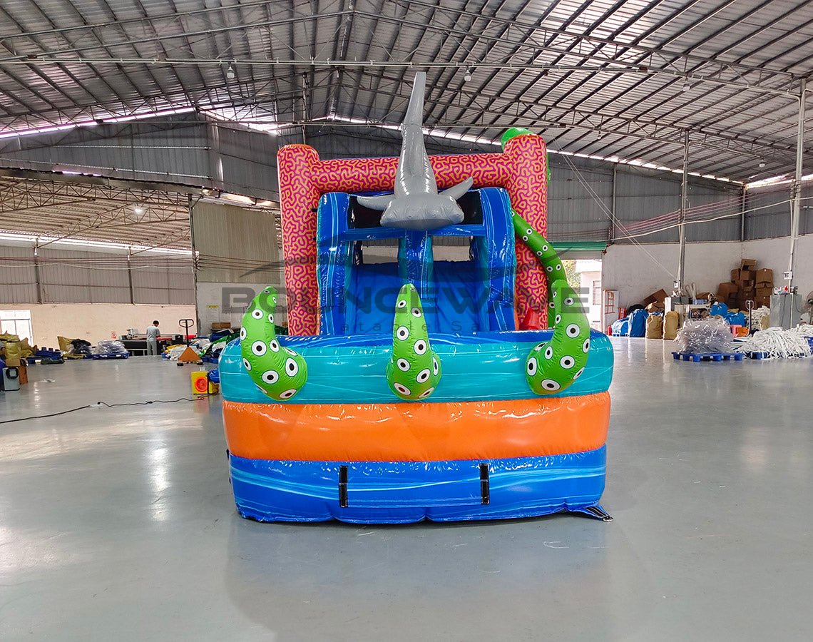 Close-up front view of Hammerhead Reef Dual Lane Commercial Inflatable Combo showing the entrance area. Features a pink coral-patterned structure with mesh windows, inflatable shark fins, and a large green octopus on top. The dual blue slides and splash pool are partially visible. Set in a warehouse with concrete flooring and various other inflatable products in the background, showcasing its commercial use context.
