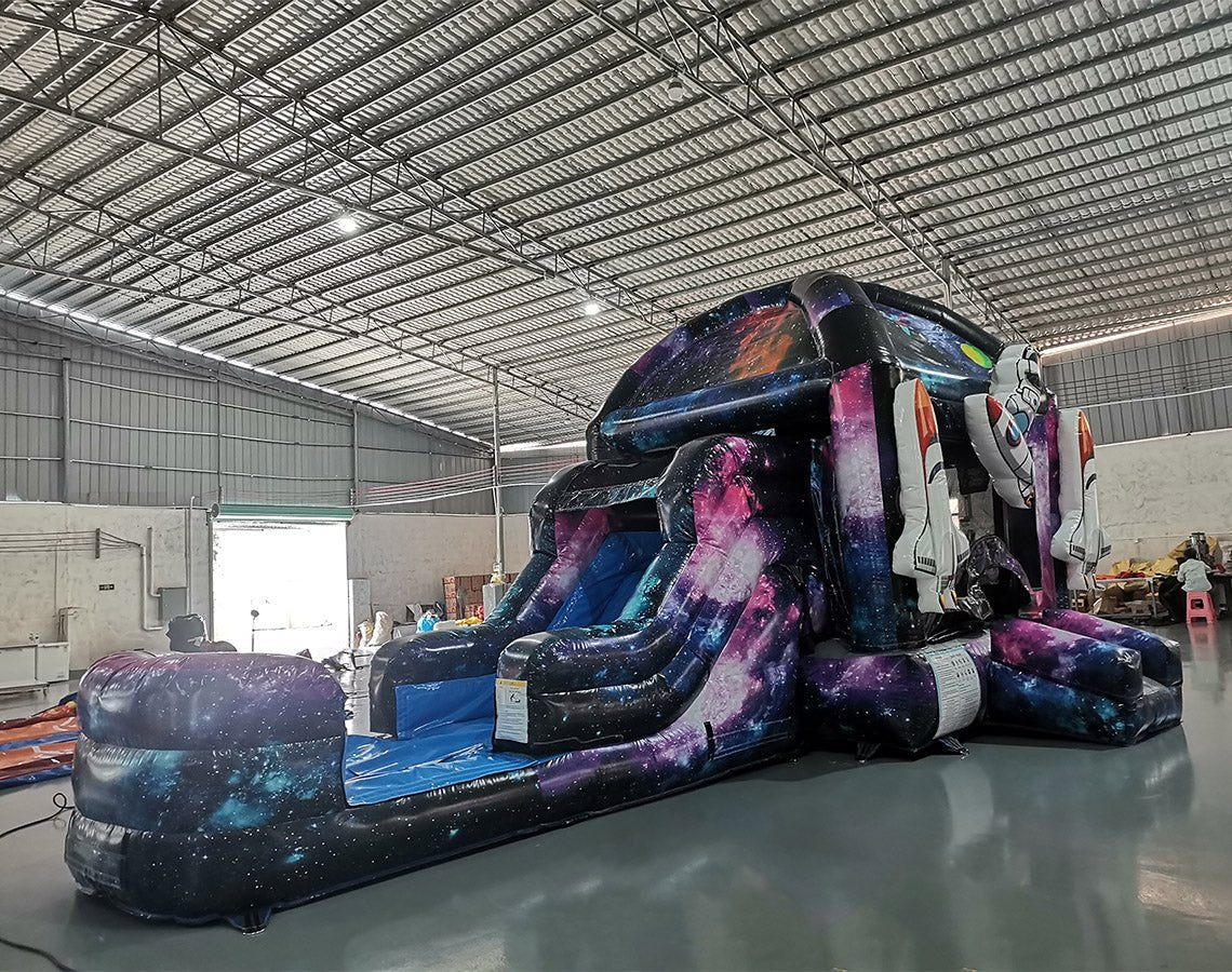 Wide-angle view of MoonWalker Commercial Econo Inflatable Wet/Dry Combo featuring a space-themed bounce house with cosmic design, attached slide, and play area. The inflatable has bright purple, blue, and pink colors with astronaut and rocket decorations. Set up in an indoor warehouse with metal roofing and other inflatable products visible.